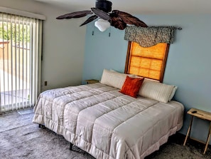 A bedroom with a large bed, a ceiling fan, a window with a patterned curtain, and a view of the outdoors.