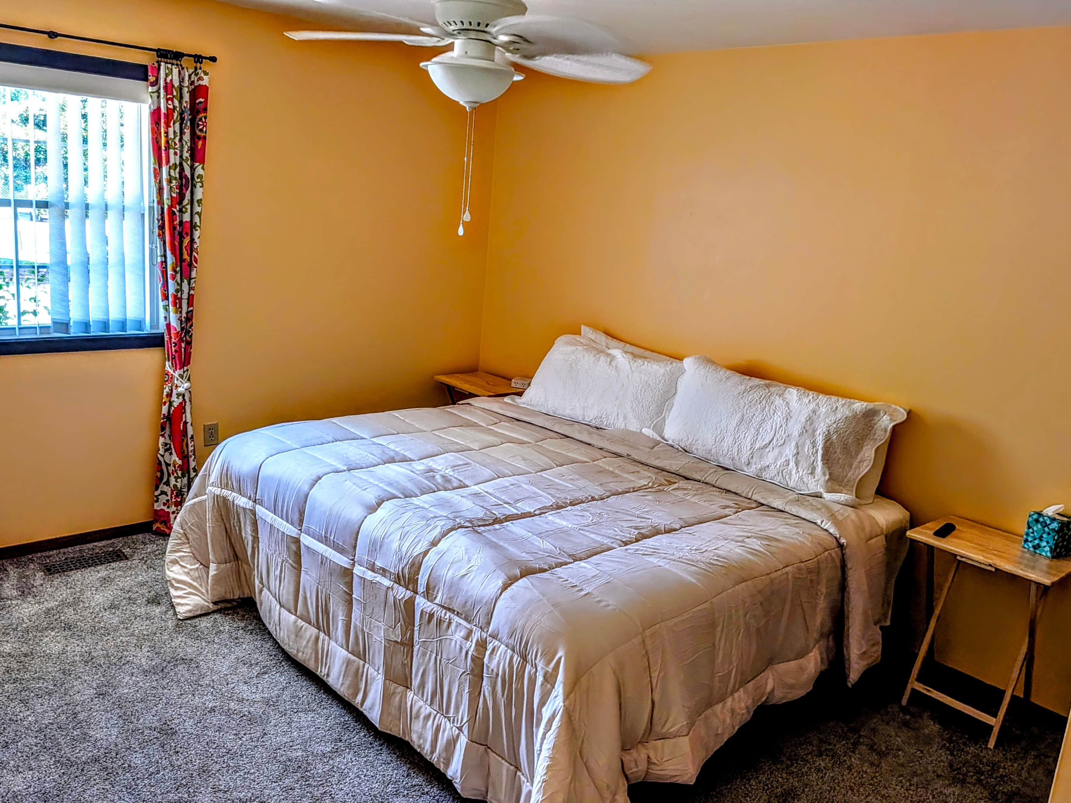 A bedroom with a bed, a nightstand, a ceiling fan, and a window with curtains.