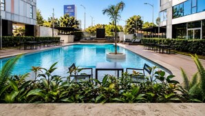 Una piscina con sillones y mesas, rodeada de vegetación y edificios modernos.