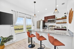 A modern kitchen with a large window, bar stools, and stainless steel appliances. - Luxe 4BR: Boardwalk + 3rd Floor Deck (Port Aransas)