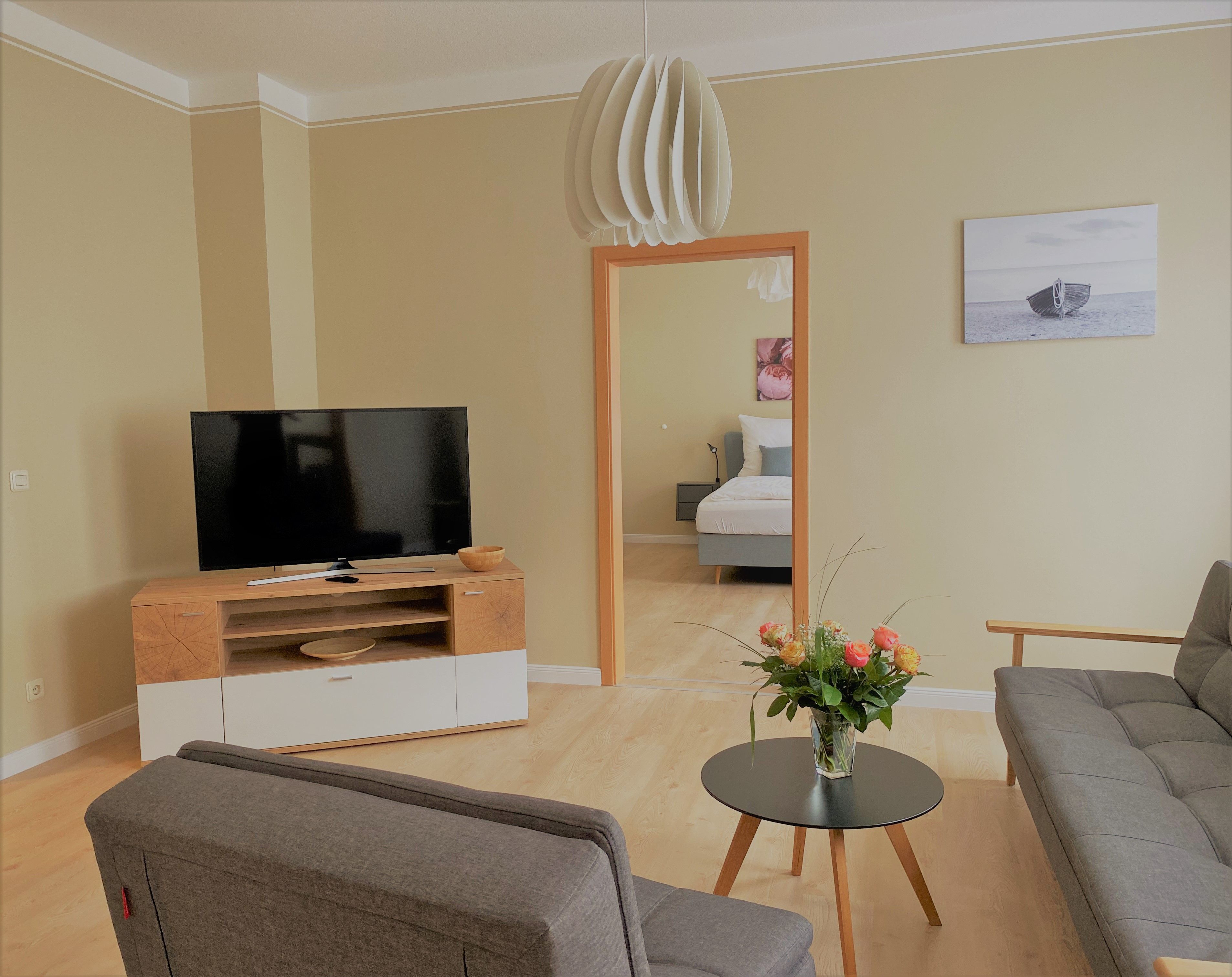 A modern living room with a flat-screen TV, a wooden entertainment unit, a grey sofa, a small round table with a vase of flowers, and a painting on the wall.