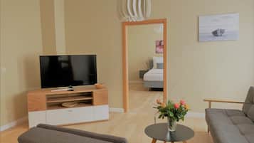 A modern living room with a flat-screen TV, a wooden entertainment unit, a grey sofa, a small round table with a vase of flowers, and a painting on the wall.