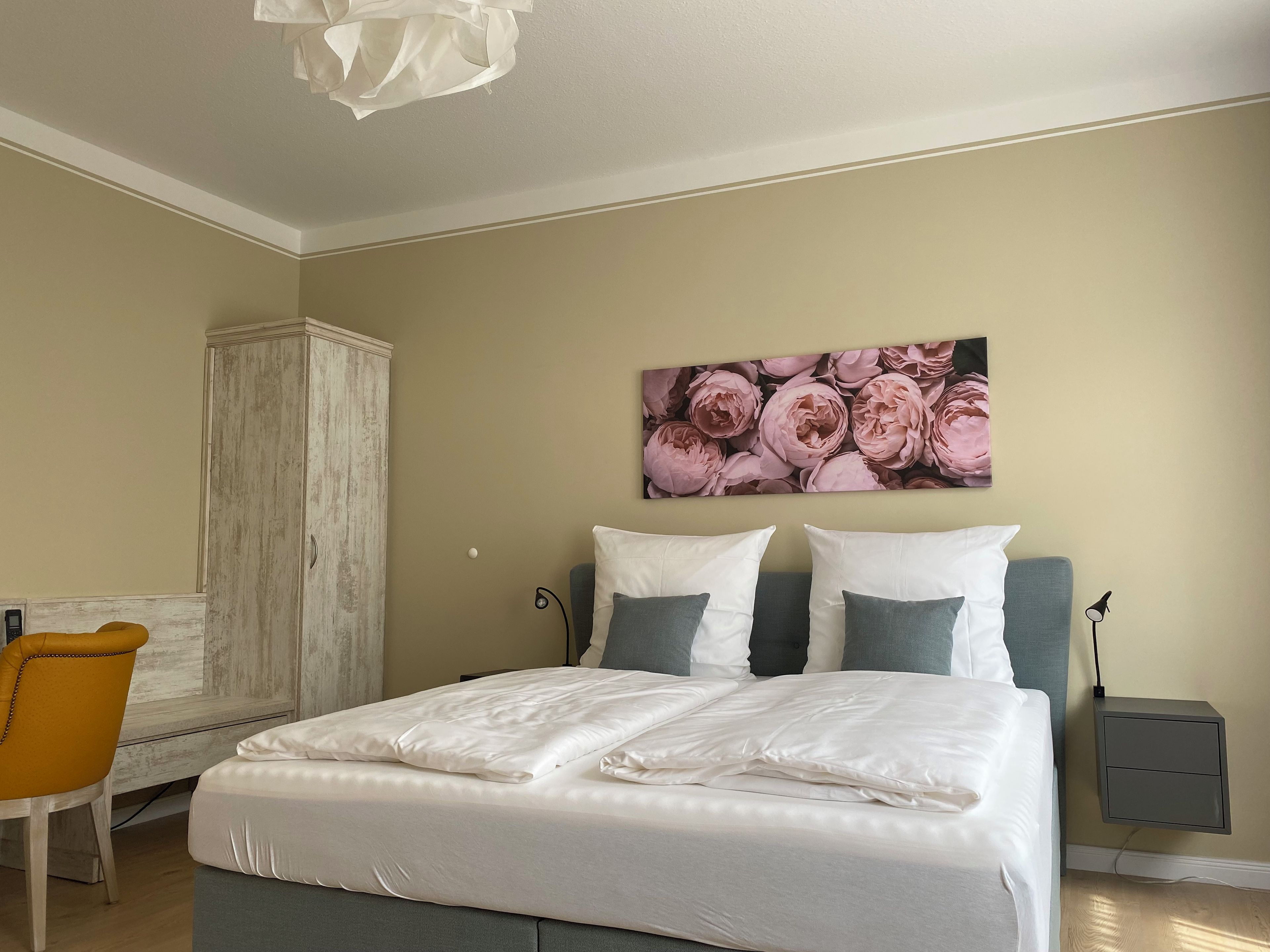 A bedroom with a bed, a nightstand, a chair, a wardrobe, and a floral artwork on the wall.
