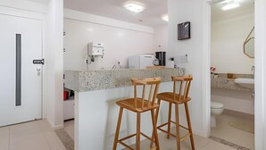 A modern kitchen with a granite countertop, two wooden bar stools, a coffee machine, and a refrigerator. - Pé de Areia Barra Ondina (Salvador)