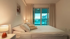 A hotel room with a bed, a nightstand, a lamp, a window with curtains, and a view of a cityscape. - Pé de Areia Barra Ondina (Salvador)