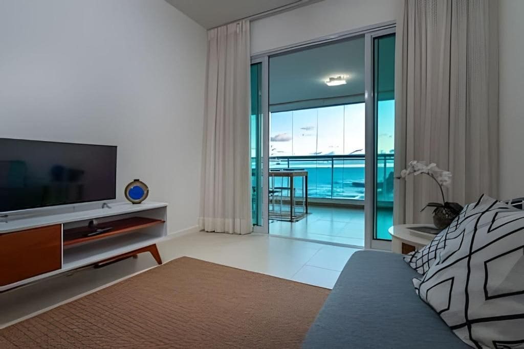 A modern hotel room with a large TV, a balcony with a view of the sea, and a bed with patterned pillows.