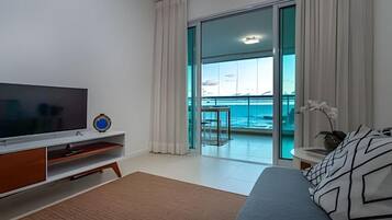 A modern hotel room with a large TV, a balcony with a view of the sea, and a bed with patterned pillows.