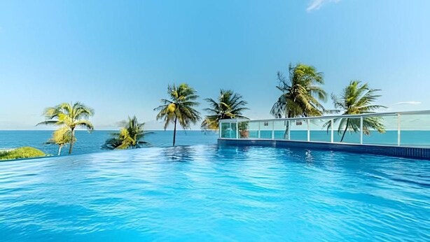 A swimming pool with clear blue water, surrounded by palm trees and a building with a glass facade. - Pé de Areia Barra Ondina (Salvador)