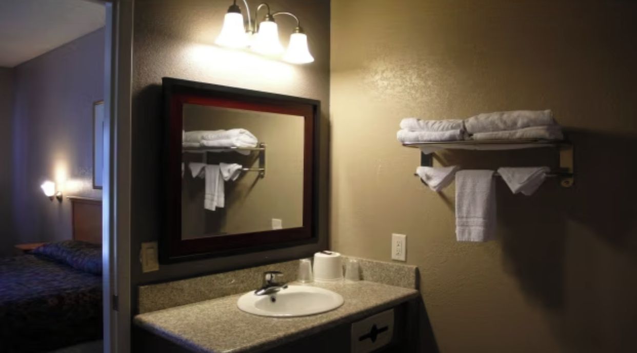 Une salle de bain d’hôtel avec un lavabo, un miroir, un porte-serviettes, et un lit visible au fond.