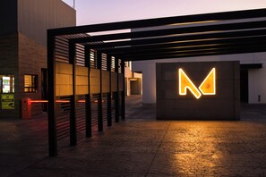 Fachada moderna de edificio con un llamativo letrero iluminado con la letra 'M', un paseo techado y pavimento de ladrillo.
