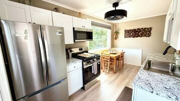 A modern kitchen with stainless steel appliances, a dining area with a wooden table and chairs, and a view of greenery outside.