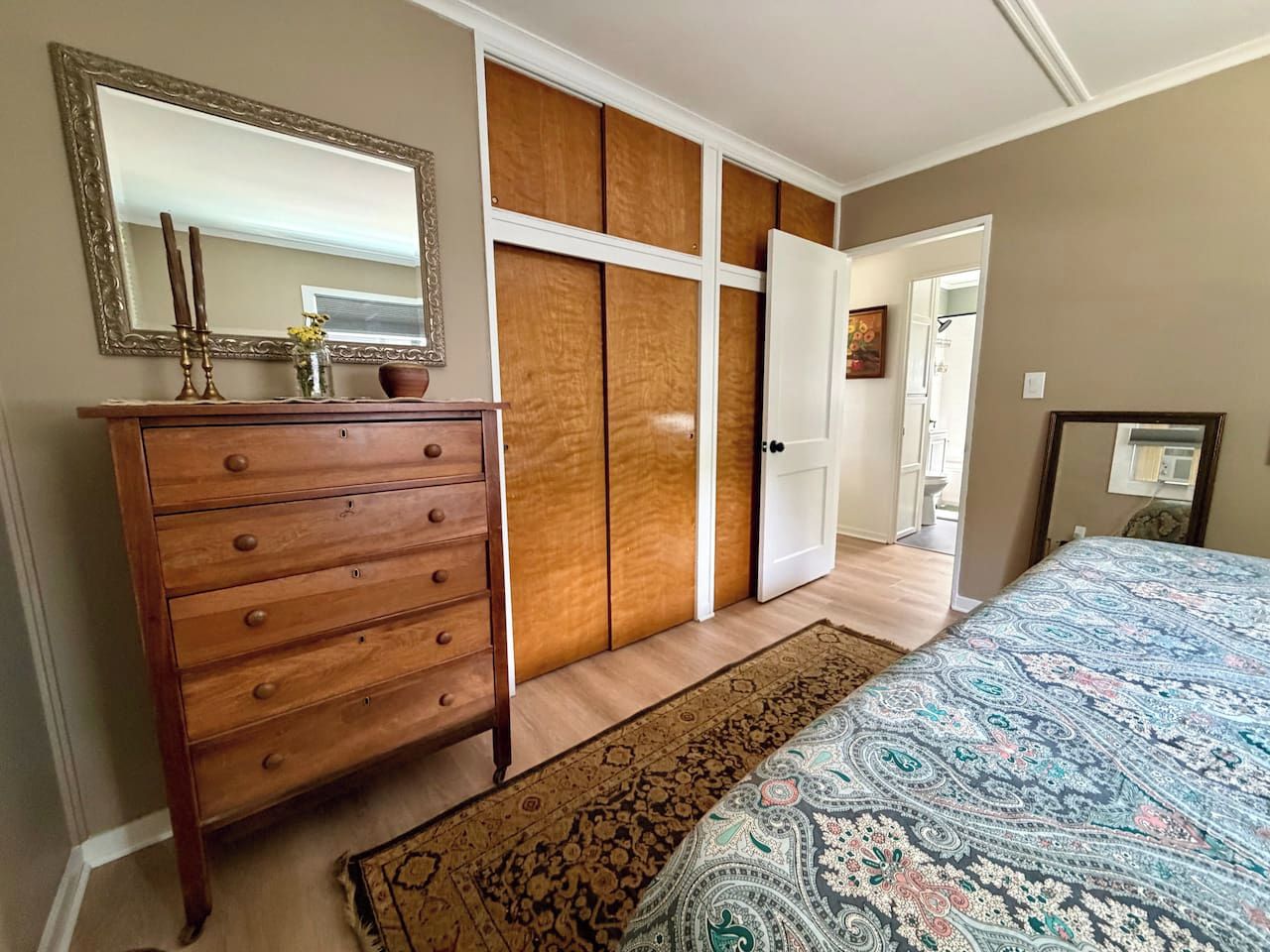 Ein Schlafzimmer mit einem Holzkoffer, einer gemusterten Bettdecke, einem großen Spiegel und einem Kleiderschrank mit Schiebetüren.
