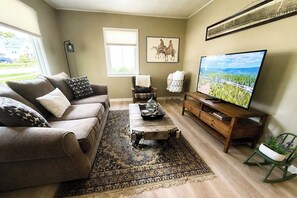 A living room with a sofa, coffee table, TV, and a view of a green landscape. - King Bed • Vintage Urban Oasis • Near MKE Airport (Milwaukee)