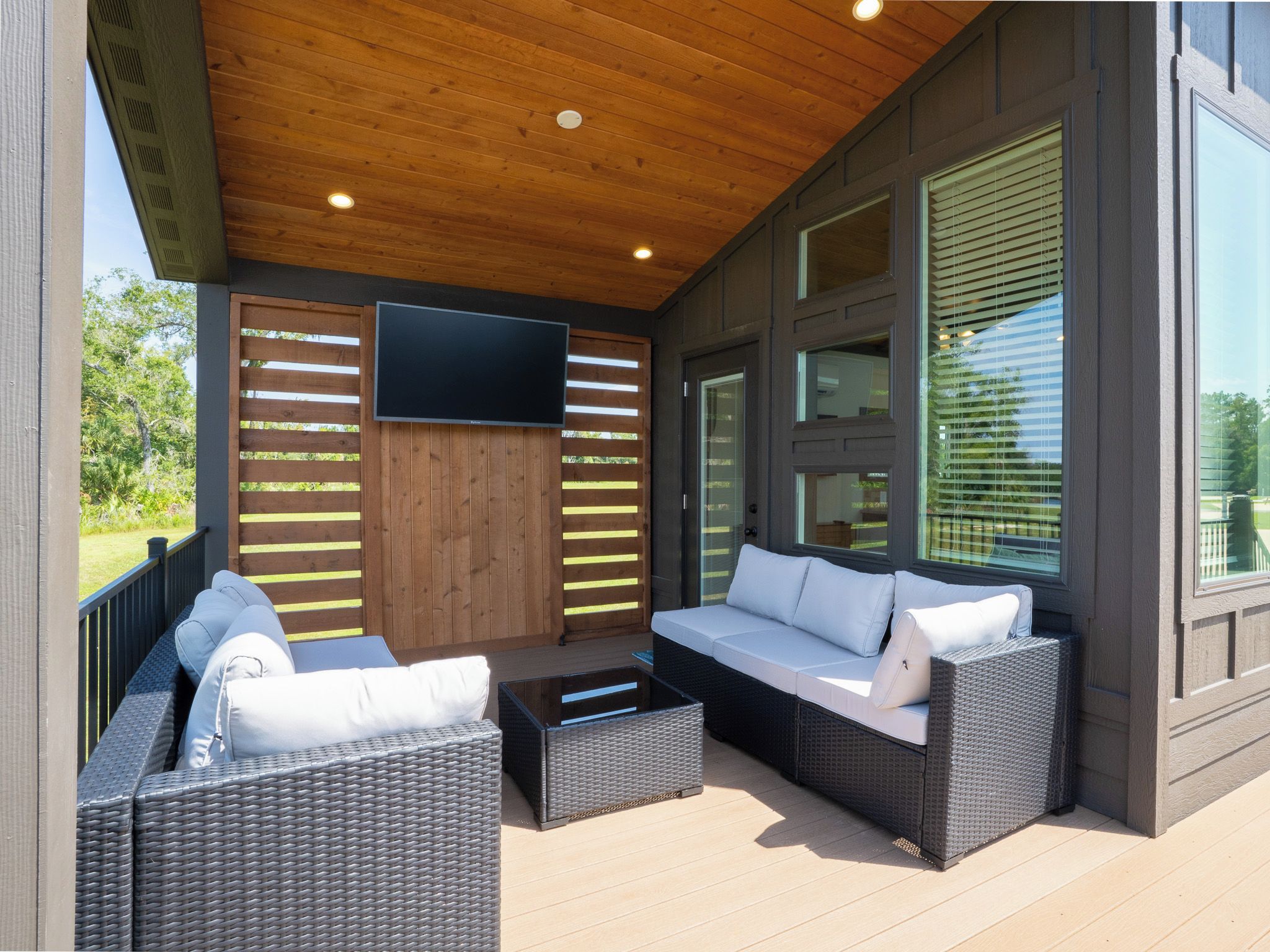A covered outdoor seating area with wicker furniture, a flat-screen TV, and large windows.