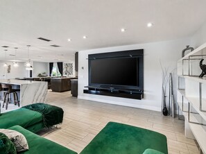 Ein modernes Wohnzimmer mit einem großen Flachbildfernseher an der Wand, einem grünen Sofa und einer Küche im Hintergrund.