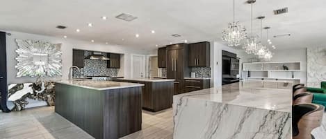 A modern kitchen with a marble island, dark wooden cabinets, and pendant lighting.
