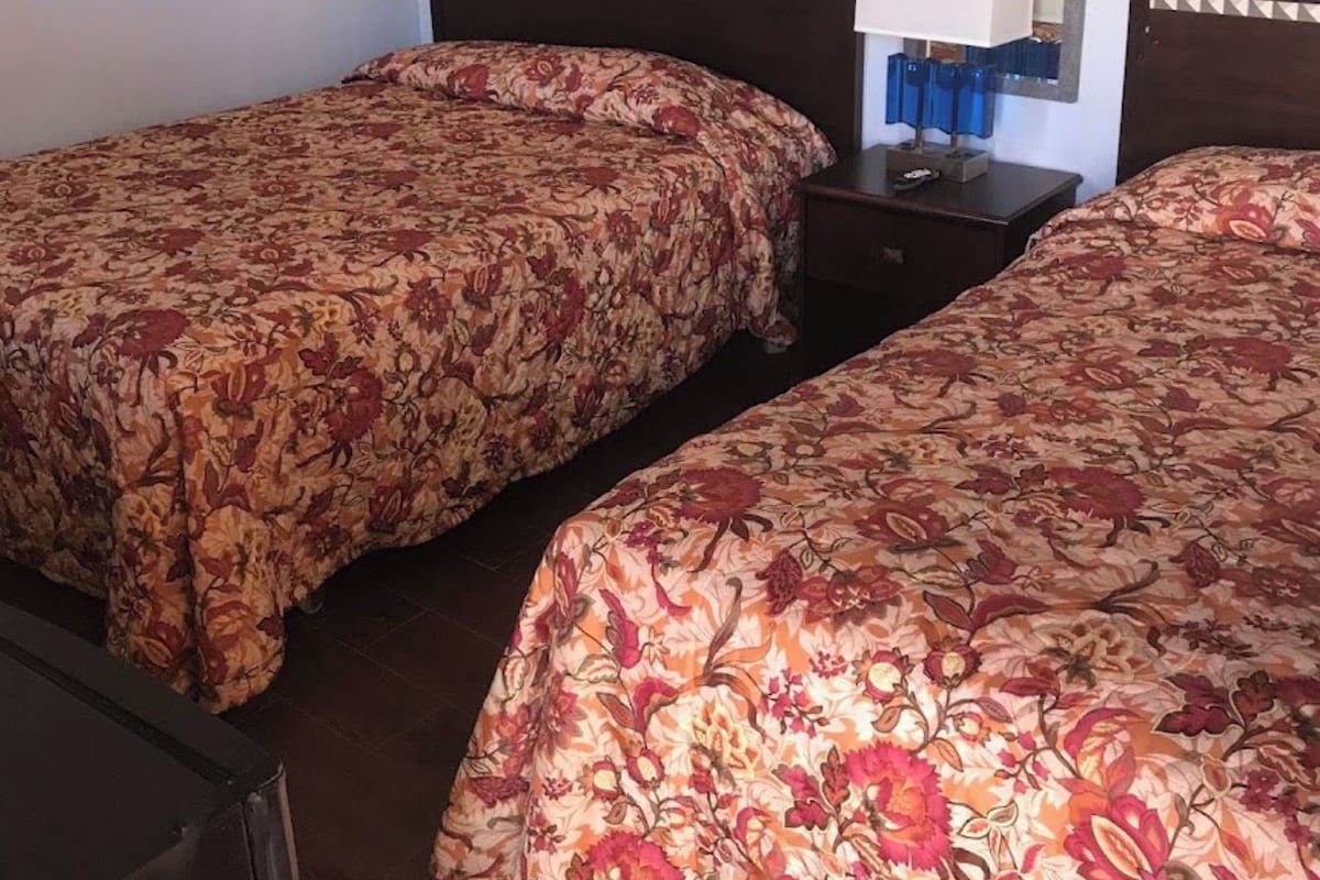 A hotel room with two single beds, a nightstand with a lamp, a mirror, and a framed picture on the wall.