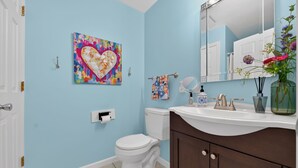 A bathroom with a white toilet, a sink with a mirror, a decorative heart painting on the wall, and a vase with flowers on the counter. - Gather at 104 - Walk to Downtown Perrysburg (Perrysburg)