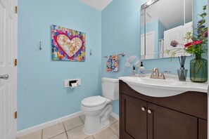 Une salle de bain avec des toilettes blanches, un lavabo avec un miroir, un tableau décoratif représentant un cœur accroché au mur, et un vase rempli de fleurs posé sur le comptoir.