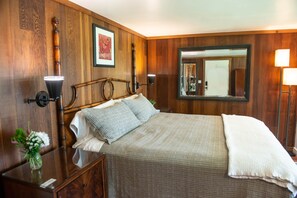 A bedroom with a wooden wall panel, a bed with pillows, a nightstand, a lamp, a mirror, and a vase with flowers. - Zen Suite at Buckhorn Cove near Mendocino w/ Ocean View, Full Kitchen, sleeps 4 (Mendocino County)
