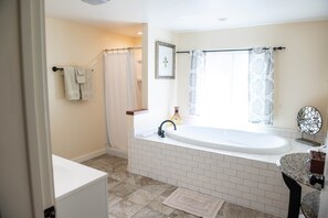Ein Badezimmer mit einer weißen Badewanne, einem Waschbecken und einem Spiegel. Ein Handtuchhalter mit Handtüchern, ein Duschvorhang und ein gerahmtes Bild sind an der Wand zu sehen.