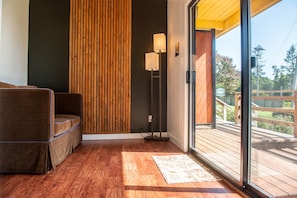 Un intérieur moderne avec un plancher en bois, un canapé, un panneau vertical en bois, une lampe sur pied et une porte en verre donnant sur une terrasse extérieure.