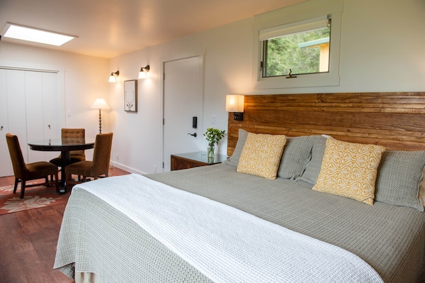 Une chambre d’hôtel avec un grand lit, une table à manger avec des chaises, une tête de lit en bois et une fenêtre donnant sur un paysage verdoyant.