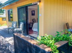 A motel with a sliding glass door entrance, a small patio area with chairs, and a bed visible inside. - Meadow Room at Buckhorn Cove near Mendocino - Cozy Gas Fireplace & EV Charger (Mendocino County)