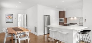 A modern kitchen with white cabinetry, a stainless steel refrigerator, a microwave, and a stove. There is a wooden dining table with white chairs and a bar stool. - Mia’s Place: Modern Pet-Friendly Geelong West Stay (Greater Geelong)