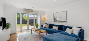 A modern living room with a blue sofa set, a flat-screen TV, and a sliding glass door leading to a patio. - Mia’s Place: Modern Pet-Friendly Geelong West Stay (Greater Geelong)