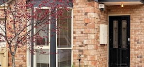 A brick building with a black door, a window with blinds, and a tree with red leaves. - Mia’s Place: Modern Pet-Friendly Geelong West Stay (Greater Geelong)