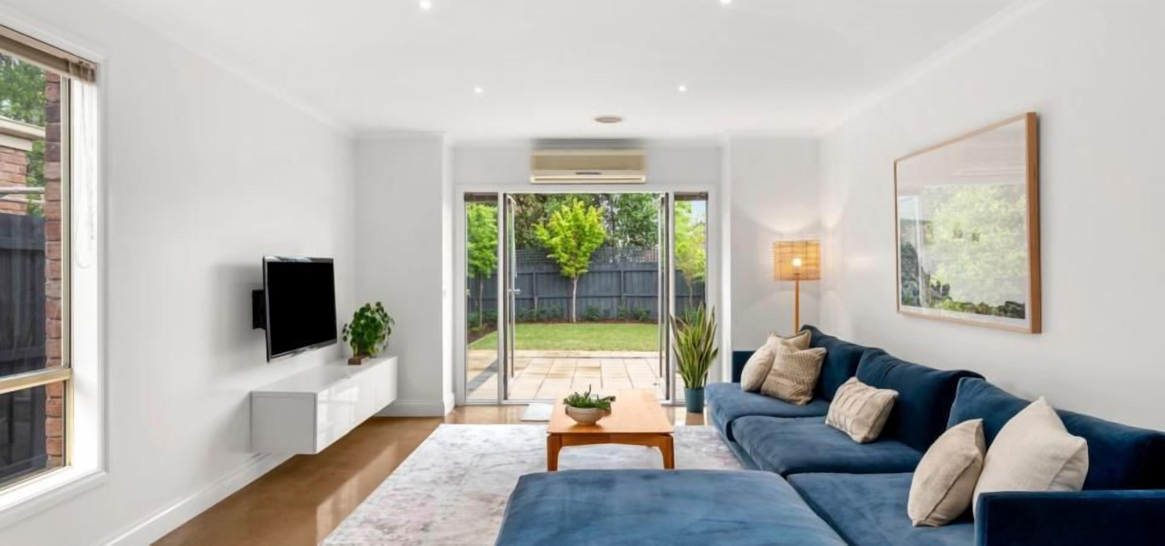A modern living room with a blue sofa, a flat-screen TV, and a large window overlooking a backyard.