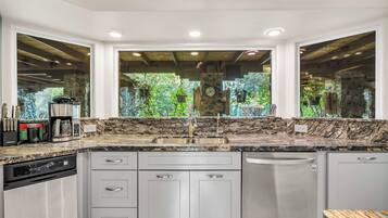 A kitchen with a granite countertop, stainless steel appliances, and a view of greenery through large windows.