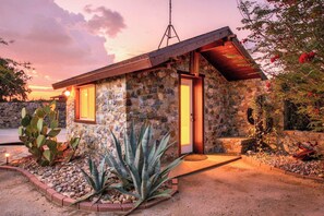 A stone house with a wooden roof, a small porch, and a cactus garden. - Desert Retreat | Hot Tub & Mountain Views (Tucson)