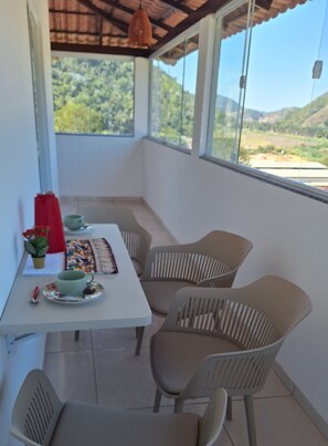 A room with a table and chairs, a window with a view of greenery and hills, and a hanging light fixture. - New apartment in Santa Teresa-ES, enclosed balcony with mountain views. (Santa Teresa)