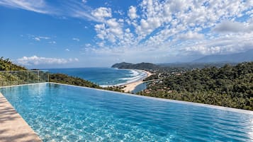 Céu azul límpido, piscina e praia com vista para o oceano e montanhas ao longe.