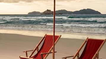 Two wooden beach chairs with red cushions and a white beach umbrella on a sandy beach with ocean waves in the background.