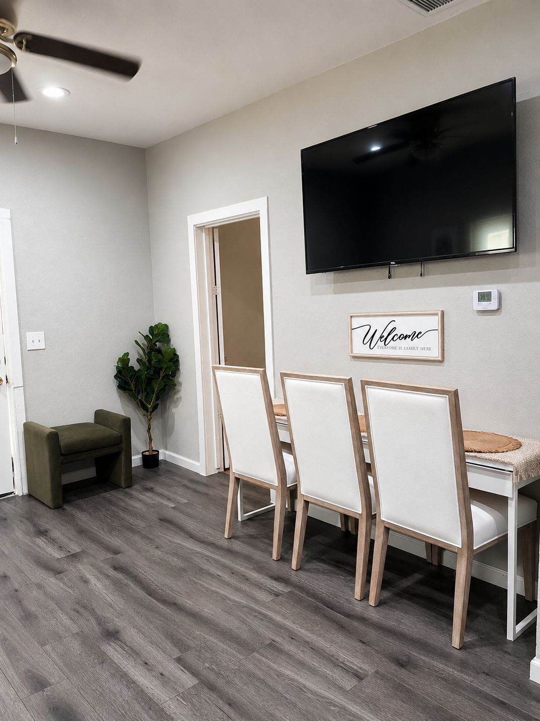 A room with a flat-screen TV mounted on the wall, a dining table with chairs, a small bench, and a potted plant.
