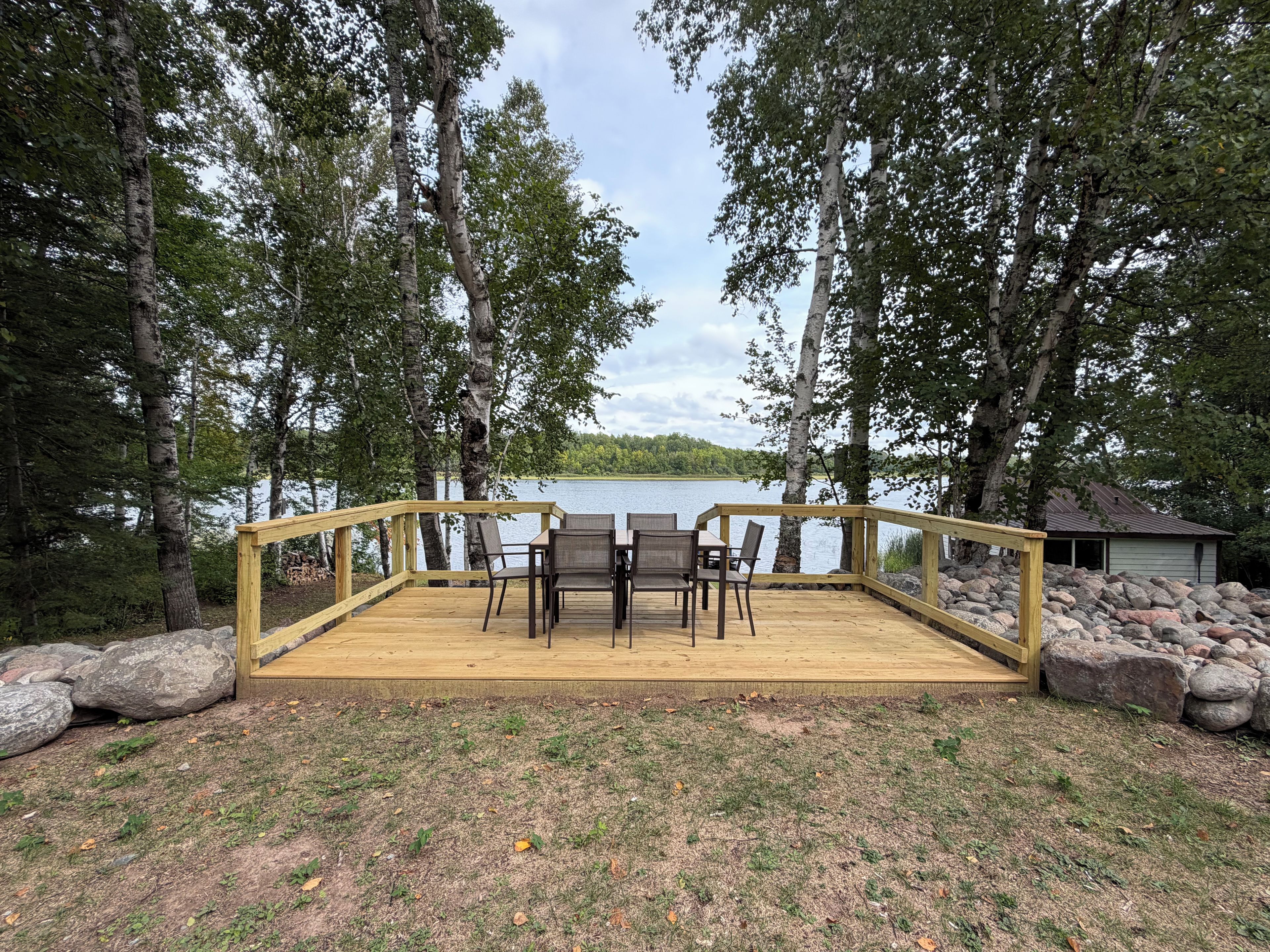 A wooden deck with a table and chairs overlooking a lake, surrounded by trees and rocks.