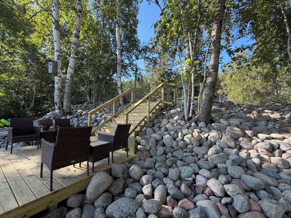 Une terrasse en bois, agrémentée de meubles en osier, et un chemin de pierre menant à un escalier en bois, le tout entouré d’arbres.