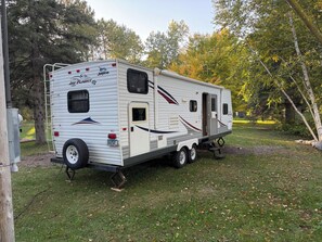 Un camping-car à l’arrêt, équipé d’une échelle, d’une porte et d’une fenêtre, garé sur une pelouse, avec des arbres en arrière-plan.