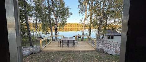 Een terras met een tafel en stoelen, uitkijkend op een meer met bomen en een blokhut.