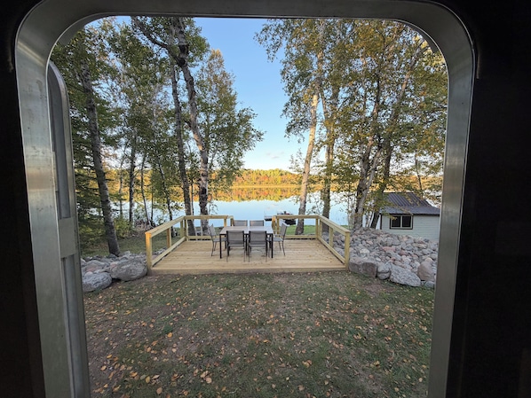 A deck with a table and chairs, overlooking a lake with trees and a cabin.