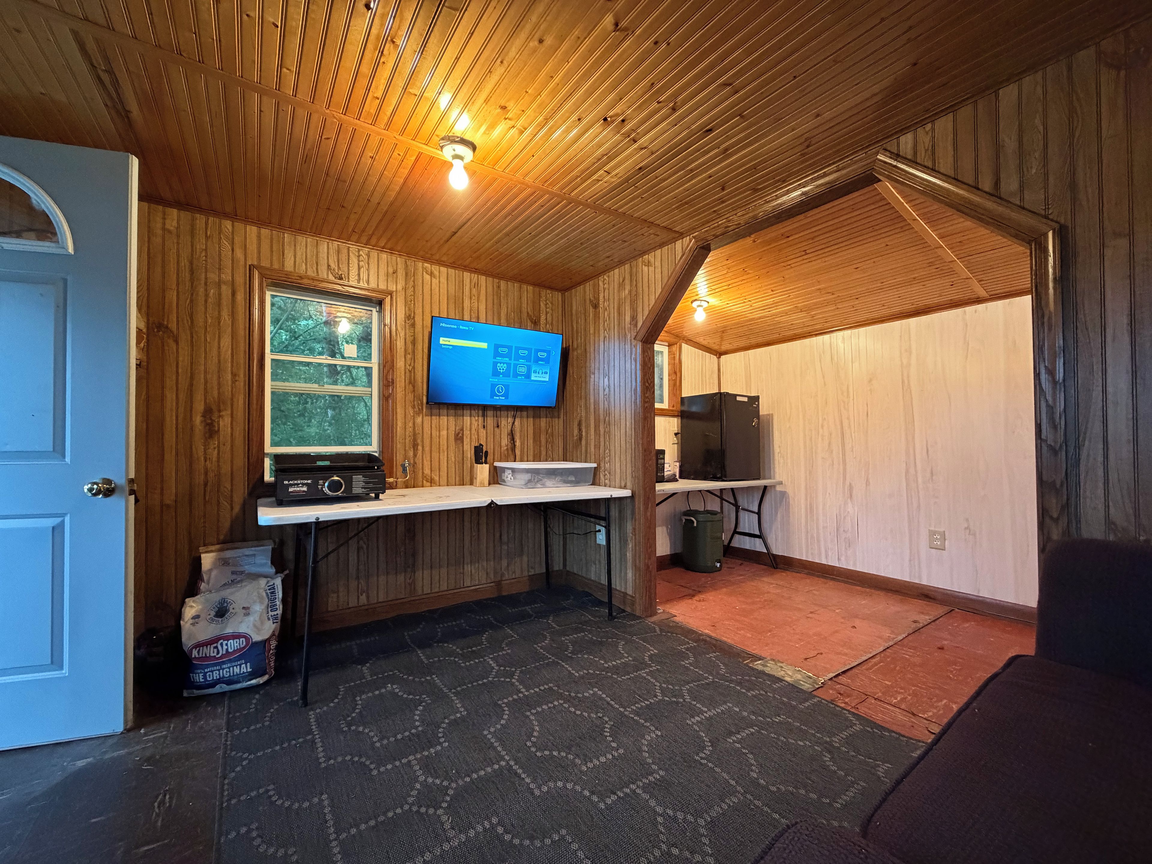 A room with wooden walls, a mounted television, a small table, a trash bin, and a door.