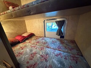Ein kleines Zimmer mit Etagenbett, einem Einzelbett mit floralem Bettbezug, einem kleinen Fenster mit Vorhängen und Blick ins Freie.