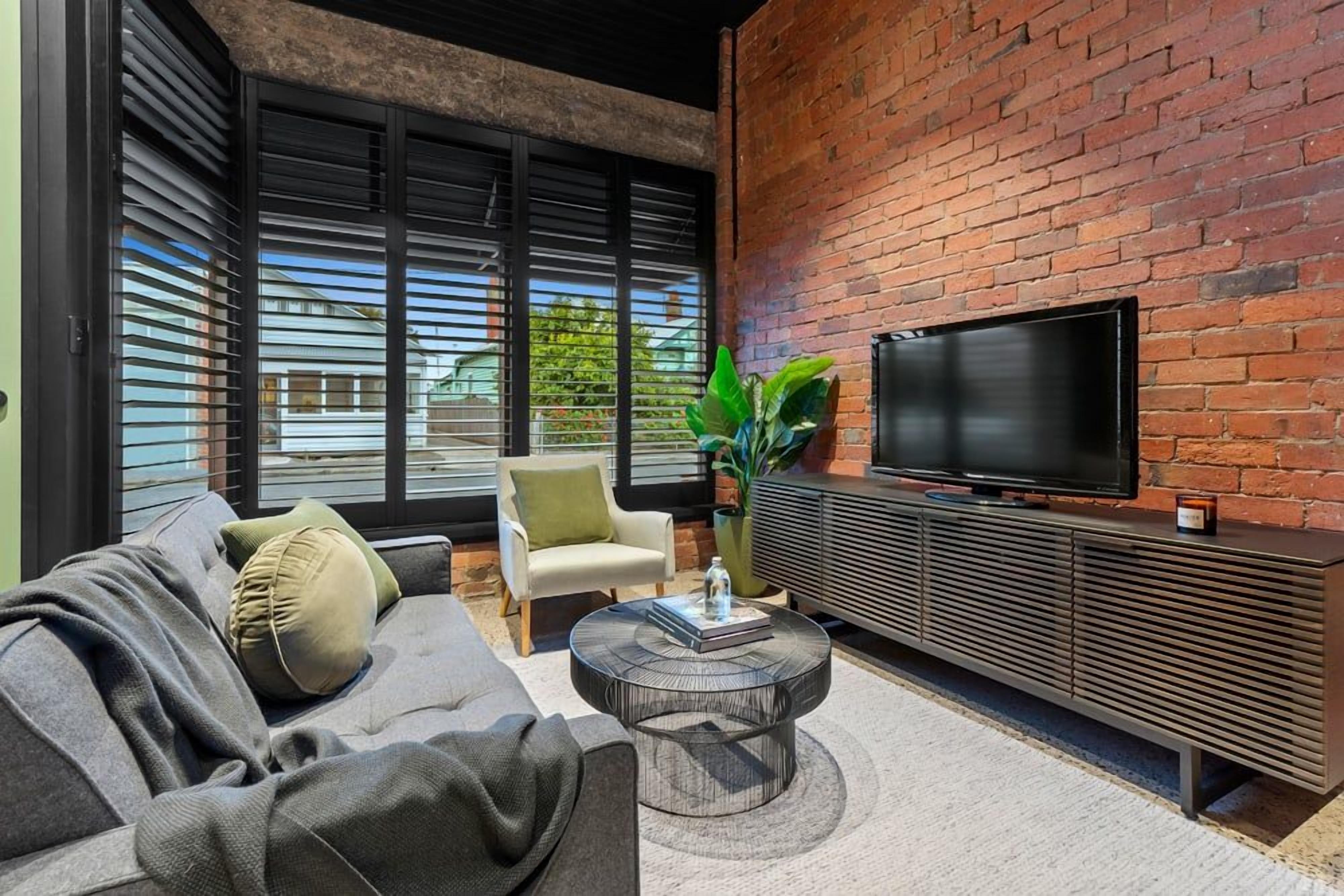 A modern living room with a sofa, armchair, and a TV on a brick wall.