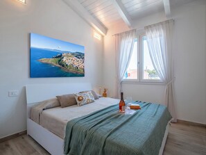 Un dormitorio con cama, un cuadro de un pueblo costero, una ventana con cortinas y una mesita de noche con una botella y un vaso.