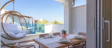 Un balcón con una mesa de madera preparada para comer, una silla colgante y vistas a edificios en la distancia.