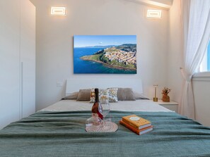 A bedroom with a bed, a painting of a coastal town, a bottle of wine, and a book. - La perla di mare 6 by Interhome (Valledoria)