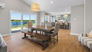 A spacious dining area with a large table and chairs, a kitchen in the background, and a view of a lake through the glass doors. - Private PEI Waterfront Escape | 5BR | Dock | Beach | Golf & Cavendish Nearby (Kensington)